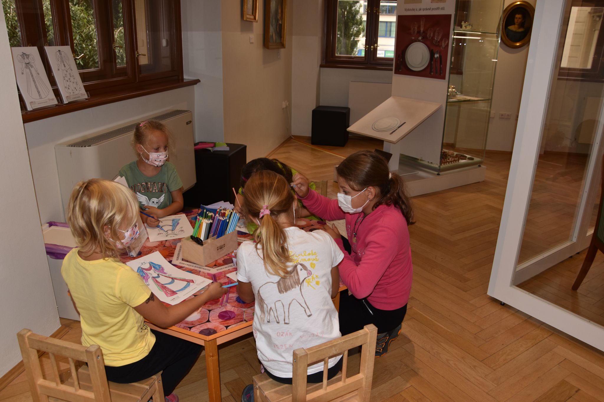 Dívky si u stolku hrají s vybarvenou vystřihovánkou oblékací panenky.